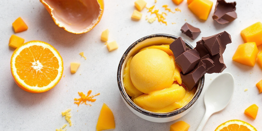 Bowl of mango tangerine sorbet topped with dark chocolate chunks, surrounded by fresh orange slices, mango pieces, and citrus zest on a light stone surface.